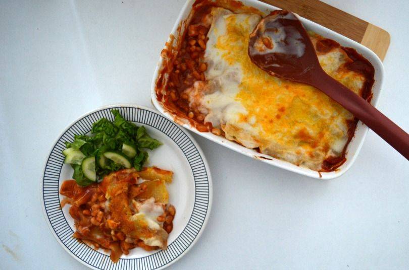 A dish of baked bean lasagne on a table with a serving spoon and a plate beside it with the lasagne on