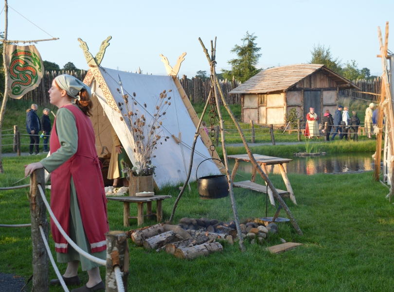 A Viking lady standing in front of a tent and a group of vikings outside a hut in the background in the Viking Village