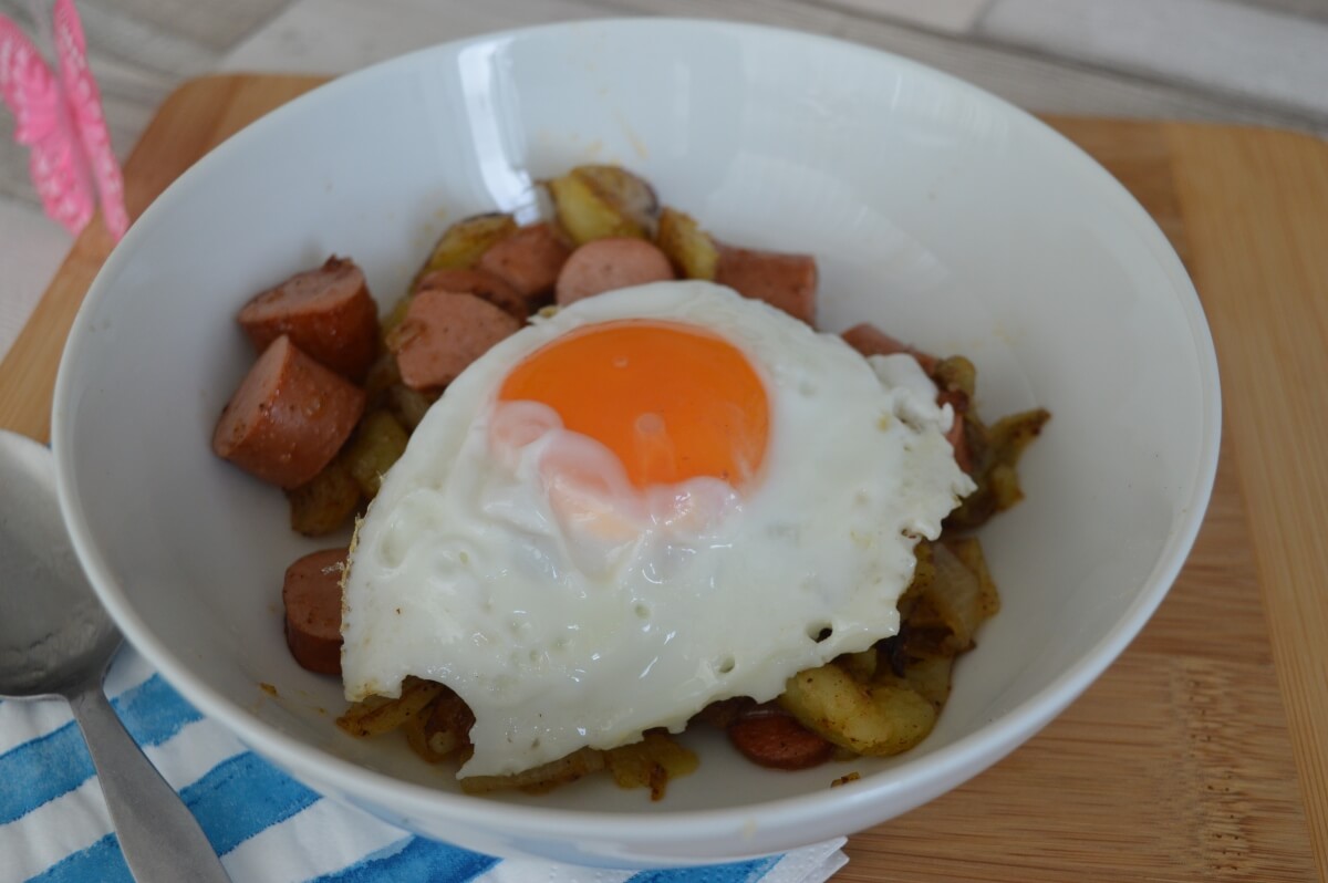 cooked chopped potatoes, onions and hot dogs in a plate with a egg on top