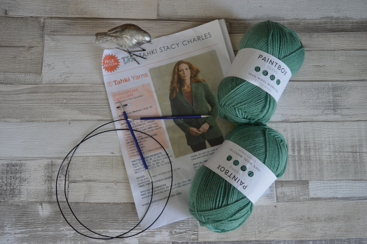 A knitting pattern, knitting needles and two balls of green wool on a wooden floor