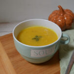 Warming spiced pumpkin soup in a pale green soup cup, garnished with pumpkin seeds beside an autumn pumpkin decoration.