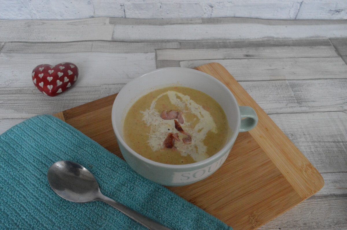 A bowl of neep and tattie (potato and swede) soup topped with bacon and cream on a chopping board with a green cloth and spoon beside it