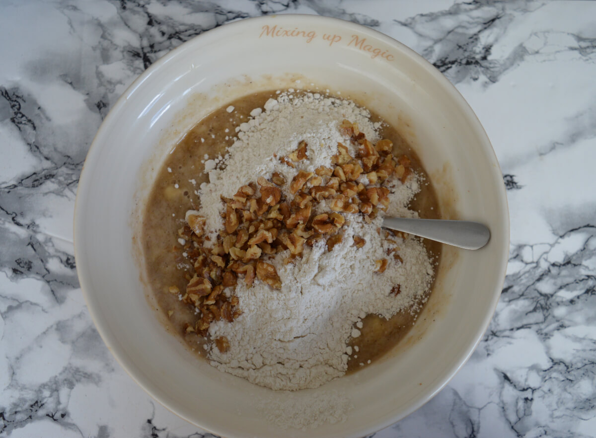 A bowl containing the ingredients for banana bread with flour and walnuts being added to the mix.