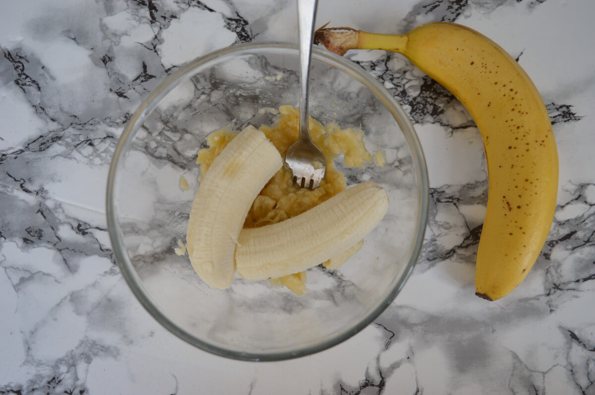 Bananas in a glass bowl being mashed with a fork. An unpeeled banana is on the counter beside the bowl.