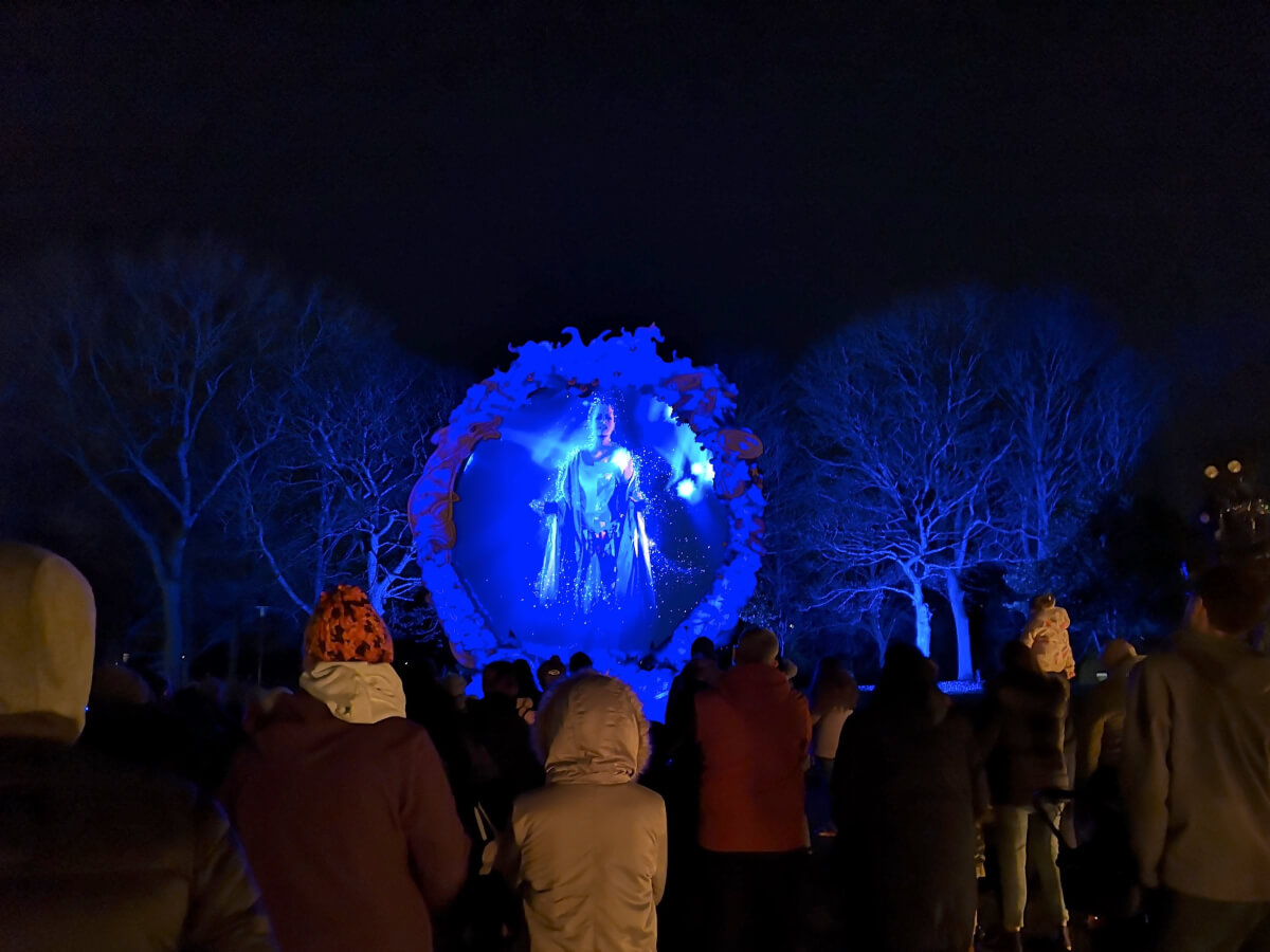 A glowing portal containing the story weaver with people standing watching