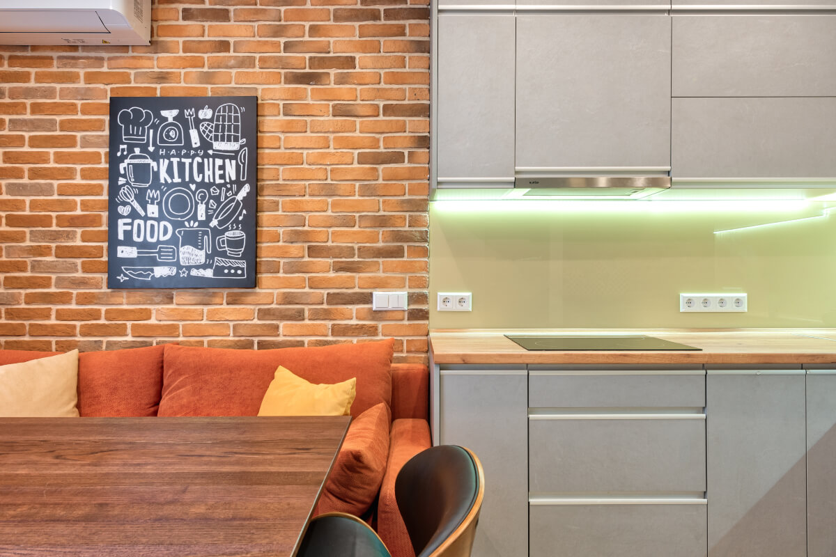 Grey kitchen cabinets lit with lights beside brown and orange sofa next to a brick wall