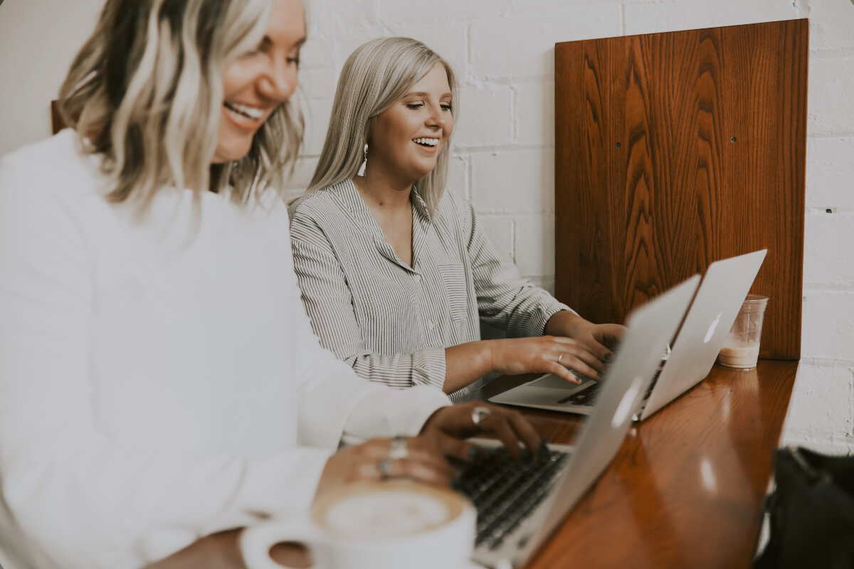 Two smiling seated women using laptops on a long brown bench